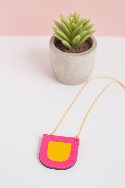 Pink and yellow bag with a chain, placed on a white surface with a potted plant in the background.