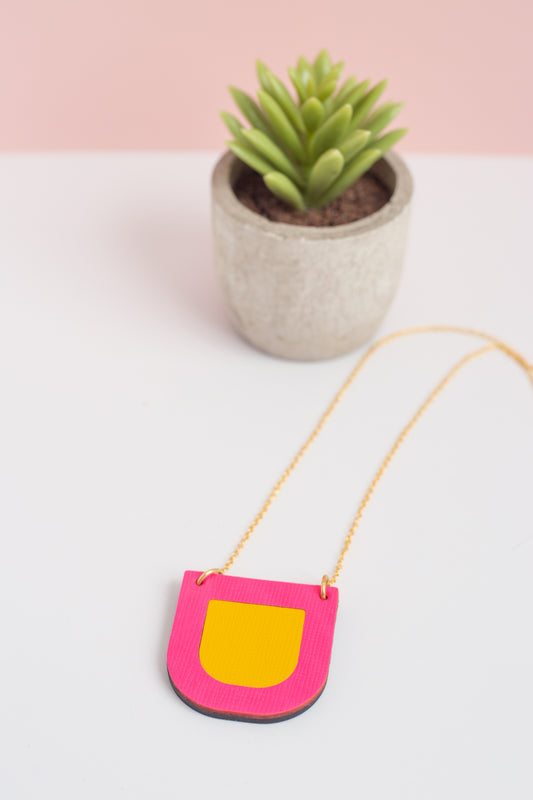 Pink and yellow bag with a chain, placed on a white surface with a potted plant in the background.