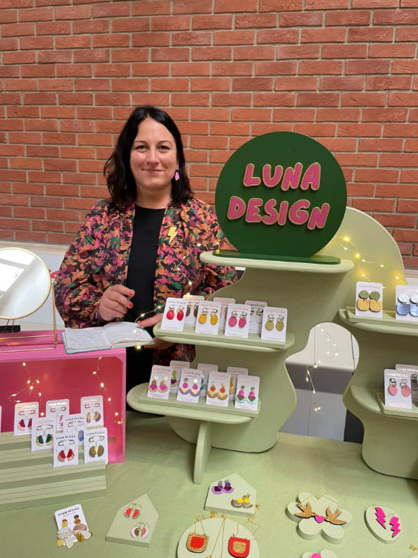 Woman standing behind a display table with 'Luna Design' branding, featuring jewelry and stationery items.