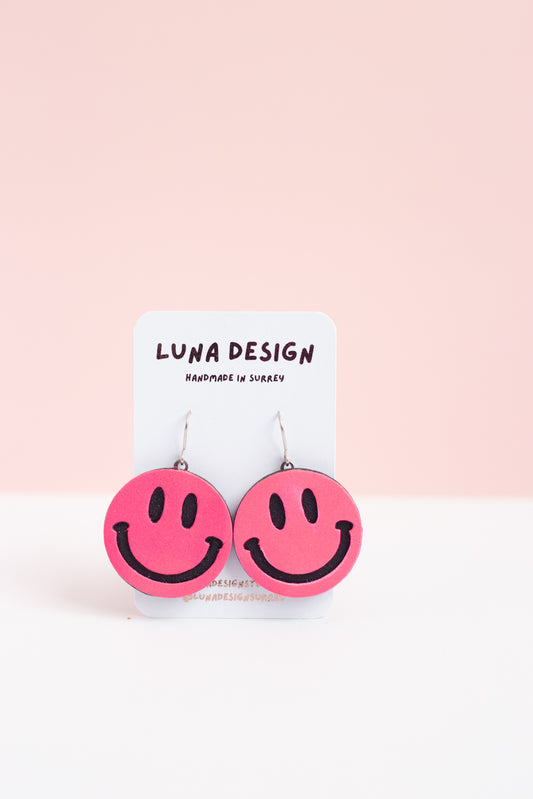 Pink smiley face earrings on a Luna Design card against a pink background