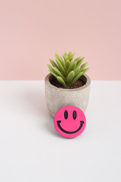 Pink smiley face object in front of a small potted plant on a white surface with a pink background