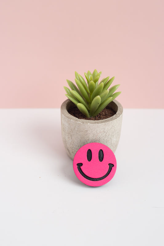 Pink smiley face object in front of a small potted plant on a white surface with a pink background