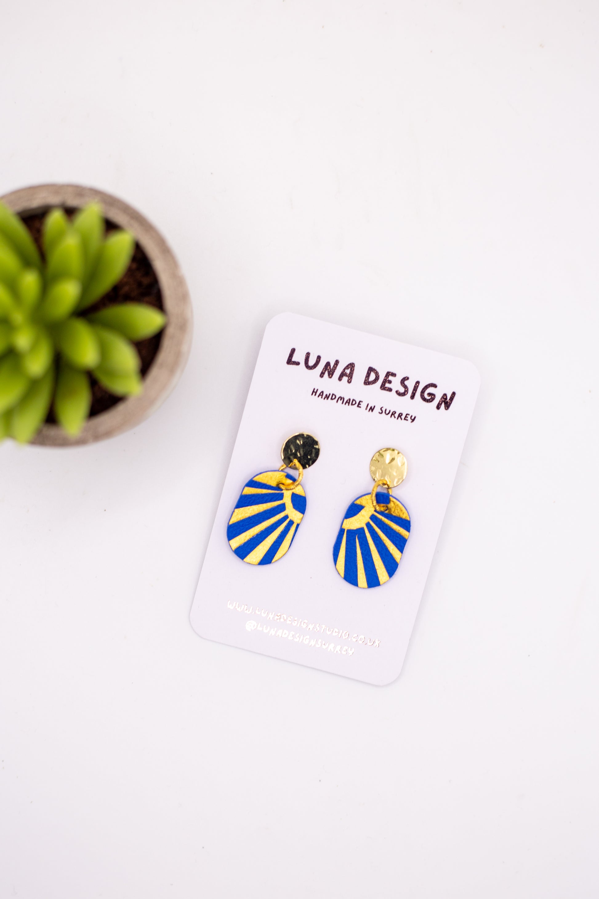 Blue and gold earrings on a Luna Design card with a plant in the background