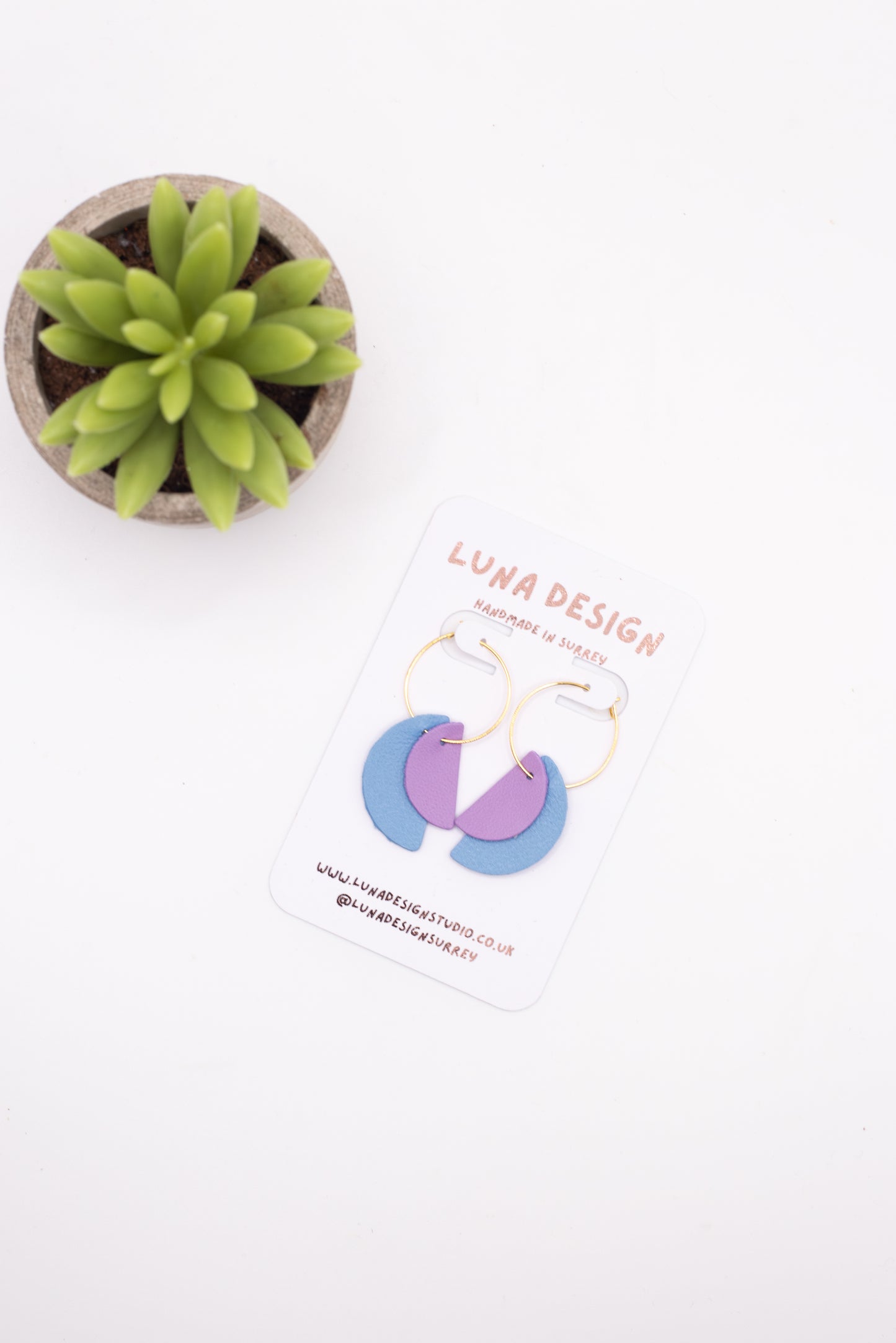 Small potted plant next to a card with earrings and 'Luna Design' branding on a white background