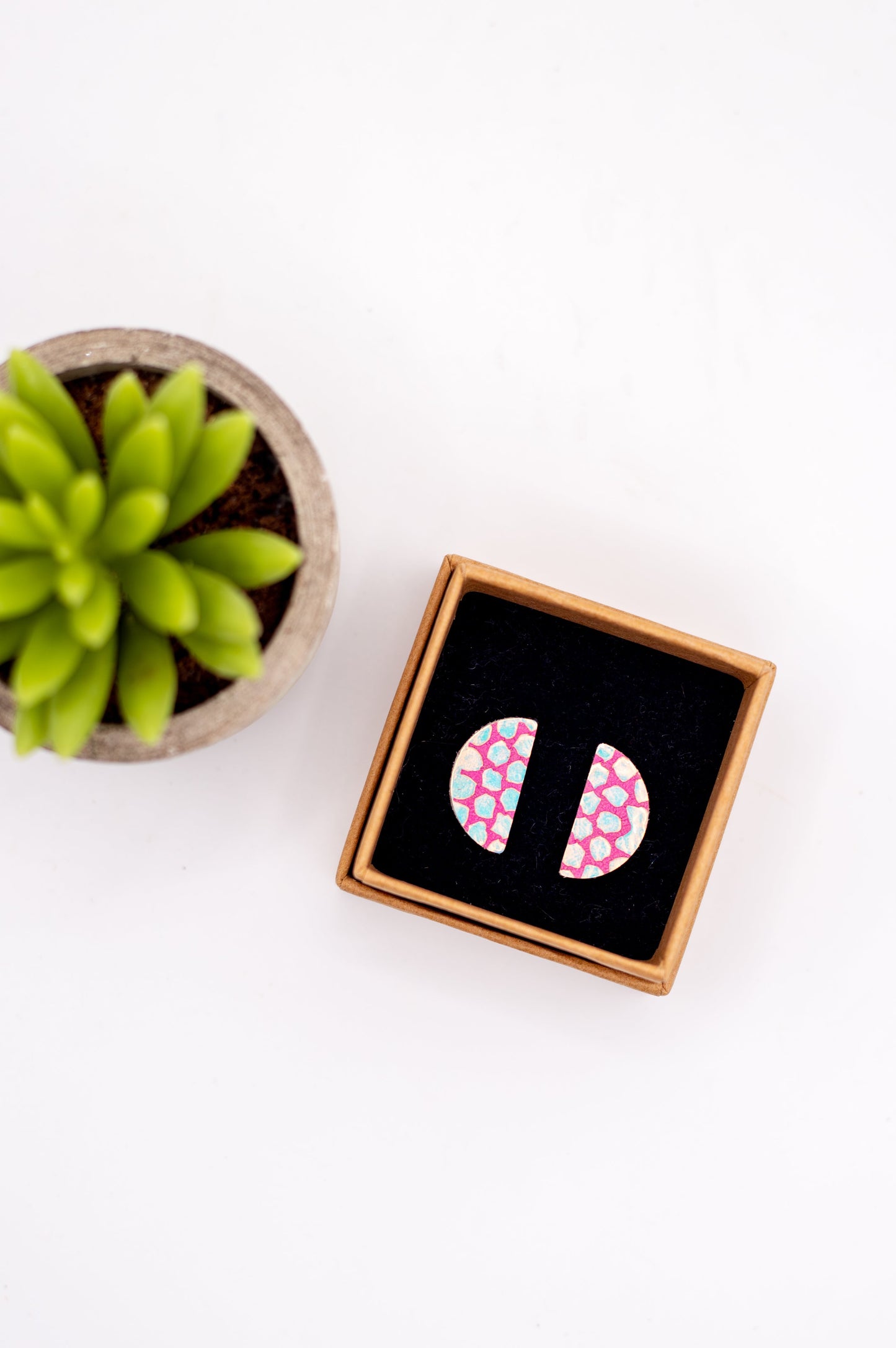 Pink and white patterned earrings in a wooden box with a succulent plant on a white background