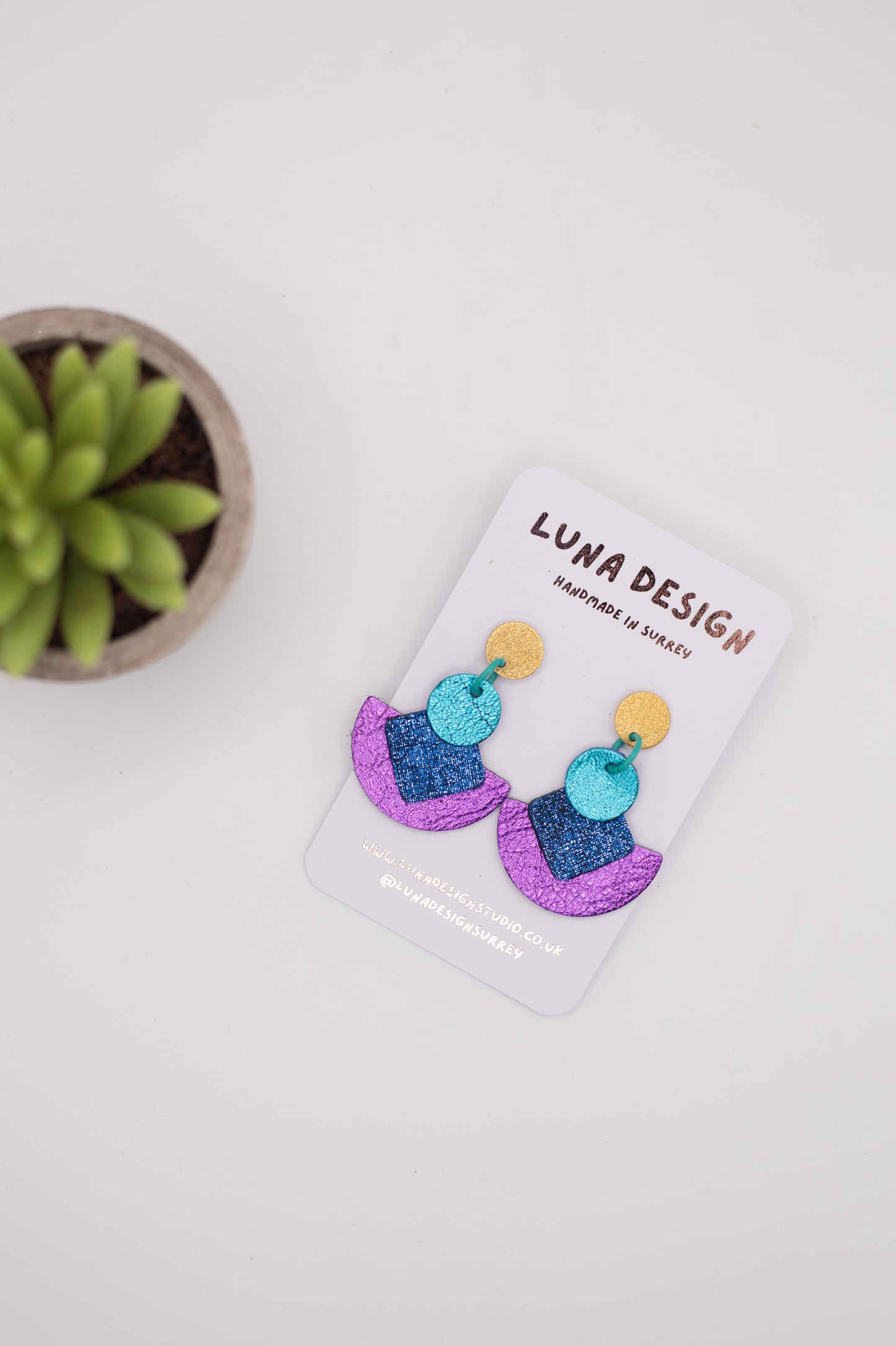 Pair of colourful Cleopatra drop earrings by Luna Design on a white branded card, featuring gold stud tops, turquoise circular links, textured dark blue squares and curved purple drops, photographed on a white background beside a small green plant.