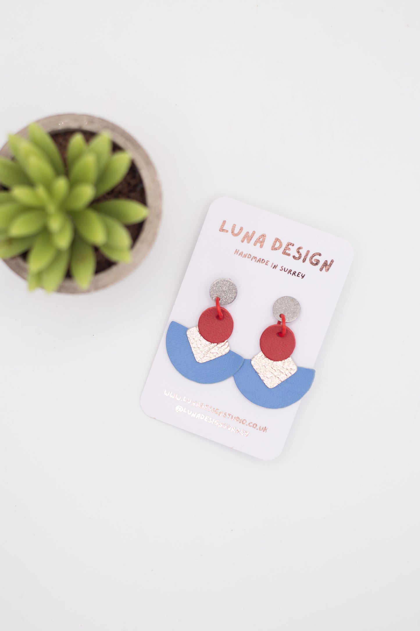 Pair of colourful statement drop earrings by Luna Design on a white branded card. The earrings feature silver stud tops, red circular links, textured pale pink squares and curved blue semi-circle drops, photographed on a white background beside a small green plant.