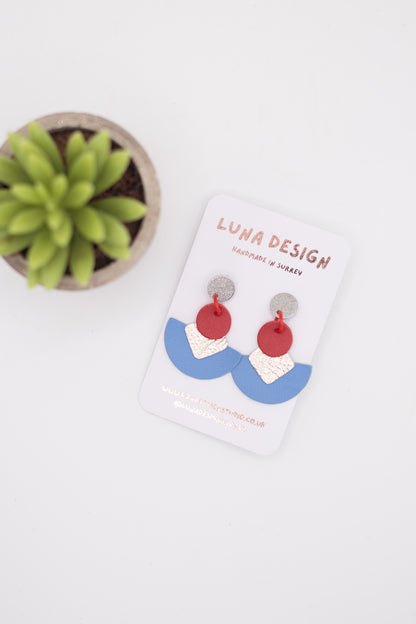 Pair of colourful statement drop earrings by Luna Design on a white branded card. The earrings feature silver stud tops, red circular links, textured pale pink squares and curved blue semi-circle drops, photographed on a white background beside a small green plant.