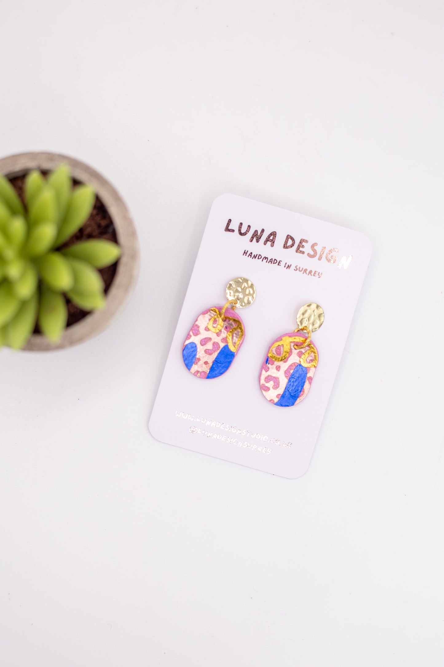 Colorful earrings on a white card with 'Luna Design' branding, next to a small plant.