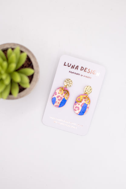 Colorful earrings on a white card with 'Luna Design' branding, next to a small plant.