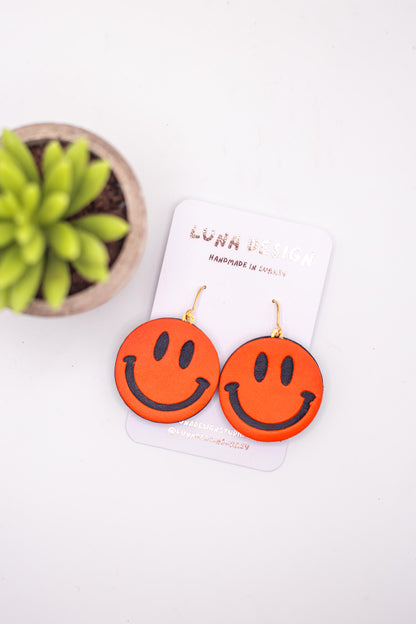 Orange smiley face earrings on a white card with a plant in the background