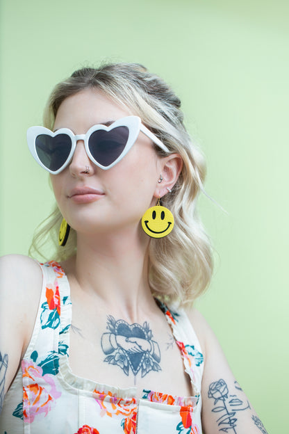 Woman wearing heart-shaped sunglasses and smiley face earrings against a light green background