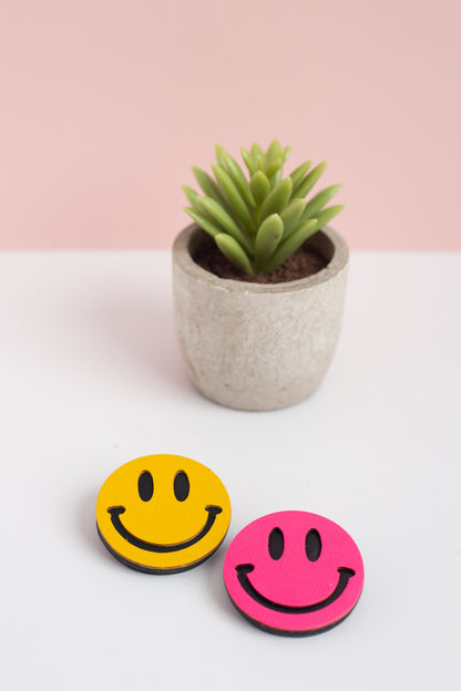 Two smiley brooch pins, one yellow and one pink, with a small potted plant on a white surface.